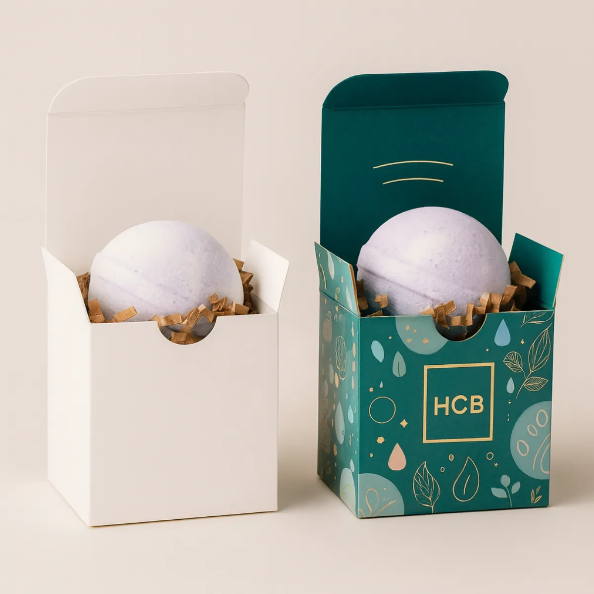 Two open cube boxes containing a single bath bomb nested in filler- one plain white and one printed in teal with an elegant gold and teal pattern-