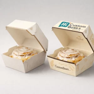 Two individual-serving hinged cardboard boxes, each containing a large, freshly glazed cinnamon roll with decorative branding.