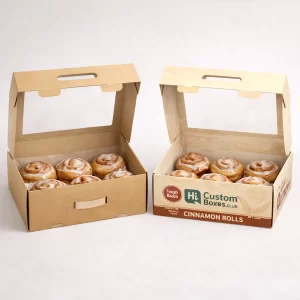 Two kraft cardboard carrier boxes with large clear windows and handles, each containing six iced cinnamon rolls.