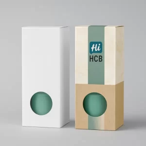 Two tall candle packaging boxes- one plain white with an open flap and the other custom printed with teal and pink candle illustrations- leaf motifs- and -Hi- branding- Both boxes feature a clear front window to display the product inside