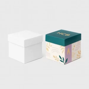 Two rigid square gift boxes with lift-off lids- one plain white and the other decorated with teal- purple- and cream accents- The designed box features the gold -HCB- logo with leaf and flame graphics